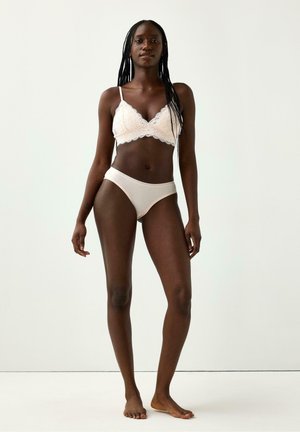 Kvinde står barfodet mod en ensfarvet baggrund iført en hvid blondebralette og matchende hvide trusser.