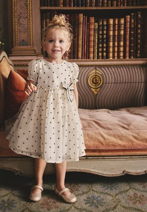 Jeune fille en robe blanche à pois et chaussures dorées sourit devant un canapé vintage avec un cadre en bois orné et de vieux livres.