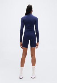 Haut ajusté à manches longues bleu marine et short assorti en tissu extensible. La tenue est complétée par des chaussures de sport blanches et des chaussettes blanches.