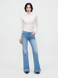 Femme aux cheveux courts et foncés portant un pull à col roulé blanc, un jean évasé bleu clair et des chaussures pointues noires, debout avec une main dans la poche.