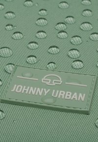 Zöld textúrával ellátott anyag vízcseppekkel és egy téglalap alakú címkével, amelyen fehér betűkkel áll a "JOHNNY URBAN" felirat, a szöveg felett egy emblémával.
