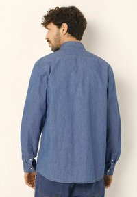 Uomo con capelli ricci e scuri, che indossa una camicia di jeans a maniche lunghe di colore azzurro chiaro e jeans blu scuro, voltato di spalle su uno sfondo semplice.