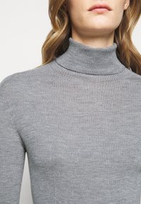 Femme portant un pull à col roulé gris côtelé, montrant les épaules, le cou et une partie du visage avec des cheveux ondulés châtain clair.
