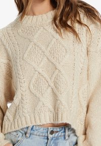 Pull en tricot ivoire avec col rond, présentant des motifs en forme de diamant et un ourlet court, fabriqué à partir d'un tissu en tricot texturé.