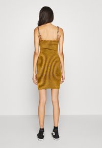 Geel en zwart gestreepte bodycon jurk met dunne bandjes en kettingdetails, gecombineerd met zwarte hoge sneakers.