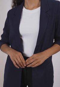 Navy blazer with notched lapels and two front pockets, paired with a white ribbed top, creating a structured, professional look.