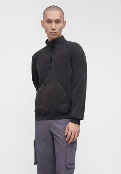 Άνδρας που φοράει μαύρο πουλόβερ με φερμουάρ ως το στήθος και τσέπη κάγκουρο, καθώς και γκρι παντελόνι cargo, στέκεται μπροστά σε απλό φόντο.