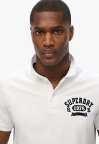 Camisa polo blanca con un logo frontal, con bordado en color azul marino. Cuello clásico y mangas cortas, fabricada en un material de algodón texturizado.