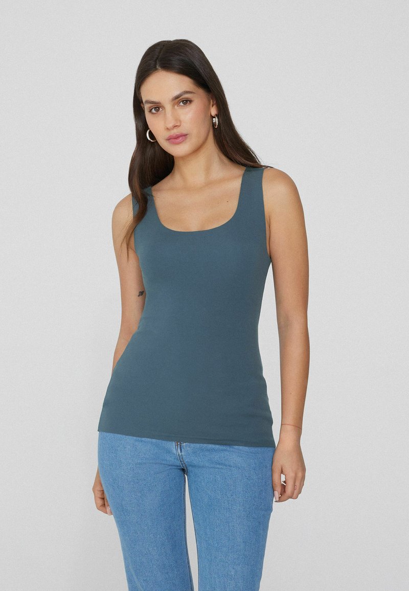 Top sin mangas para mujer en un color verde azulado oscuro, hecho de una tela suave, con escote redondo y silueta ajustada, combinado con jeans de un azul claro.