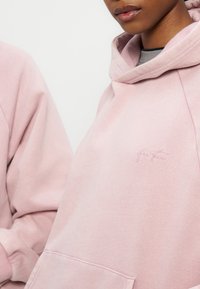Posnetek od blizu osebe, ki nosi pastelno roza pulover s kapuco z diskretnim izvezenim logotipom na prsih, pulover ima sprednji žep in kapuco.