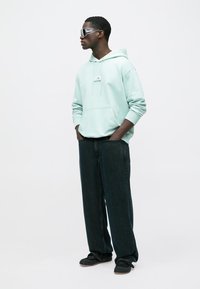 Sweat à capuche vert menthe clair avec une poche avant, associé à un jean en denim foncé ample. Le modèle porte des lunettes de soleil réfléchissantes et des chaussures noires faciles à enfiler.