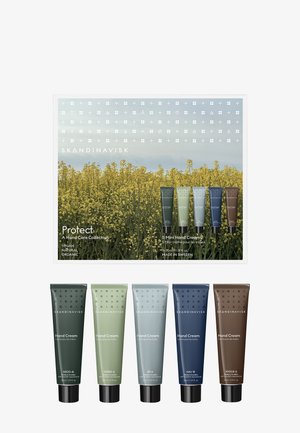 Ensemble de cinq tubes de crème pour les mains Skandinavisk en vert, vert clair, gris, bleu marine et marron, présentés avec un emballage à thème nature et des fleurs jaunes.