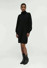Vestido de suéter preto tricotado, com decote alto, mangas balão longas e corte relaxado, combinado com botas de tornozelo pretas.