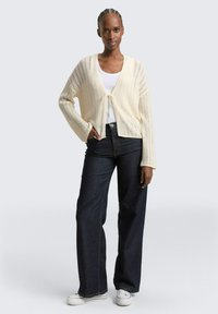 Vrouw met gevlochten haar die een crèmekleurig opengebreid vest draagt over een wit shirt, donkere wijde jeans en witte sneakers, staand met haar hand in haar zak.