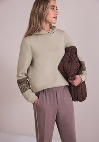 Femme portant un sweat à capuche beige avec des accents imprimés léopard sur les manches et un pantalon mauve, tenant une veste matelassée marron sur un fond uni.