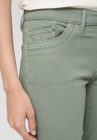 Zielone jeansy bawełniane mają szczegółowe przeszycia, przednie kieszenie i dopasowany krój. Materiał ma gładką fakturę z lekkim połyskiem.
