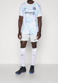 Mies jalkapalloilija, joka pukeutuu valkoiseen ja siniseen kuviolliseen pelipaitaan, valkoisiinshortseihin, valkoisiin sukkiin ja mustiin nappulakenkiin, seisoo tasaisella taustalla.