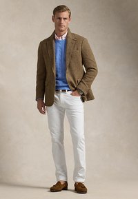 Homme portant un blazer en tweed marron, un pull en maille torsadée bleu, une chemise rose clair, un pantalon blanc, des mocassins marron et une montre, debout devant un fond neutre.