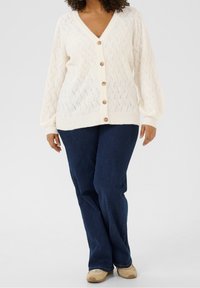 Lichtcrème gebreide cardigan met een V-hals, knopenfront en gestructureerd zigzagpatroon, gedragen met donkerblauwe denimbroek en beige schoenen.