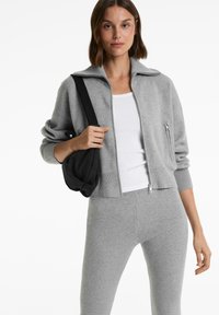 Een vrouw in een lichtgrijze zip-up jack, bijpassende legging, witte tanktop en met een zwarte schoudertas.