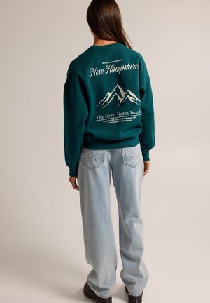 Person mit langen braunen Haaren, die einen dunkelgrünen Pullover mit Bergmotiv und hellblaue Jeans trägt, steht vor einem schlichten Hintergrund.