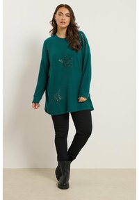 Pull en tricot turquoise avec des accents d'étoiles en sequins, associé à un jean slim noir et des bottes noires épaisses. Encolure ronde simple, manches longues.