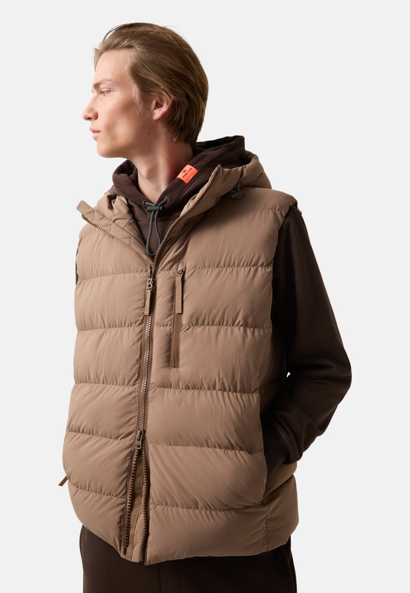 Braune Steppweste mit Reißverschluss vorne und zwei Seitentaschen. Verfügt über eine Kapuze und eine horizontale Steppung, getragen über einem braunen Hoodie.
