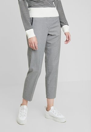 Personne portant un pantalon court à carreaux noir et blanc, un haut à manches longues rayé noir et blanc, et des baskets blanches, debout sur un fond uni.