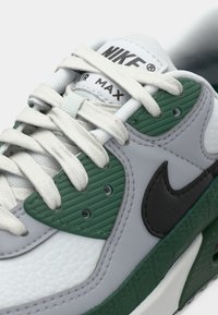 Nike Air Max kedas ar baltu ādas augšdaļu, zaļām tekstūrētām akcentiem, melnu logotipu un pelēkiem pārklājumiem. Iekļauj standarta saites un mīkstinātu zolīti.