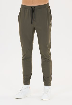 Mann trägt olivgrüne Slim-Fit-Jogginghosen mit schwarzer Kordel und weißen Sneakers vor einem einfarbigen Hintergrund.