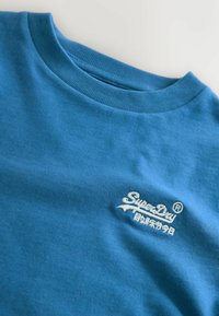 Modrá bavlnená tričko s okrúhlym výstrihom a ribbovaným lemom. Na hrudi sa nachádza biele vyšívané logo "SuperDry". Hladká textúra.