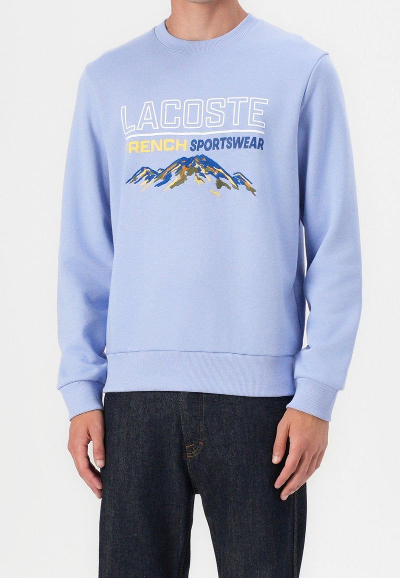 Sweat-shirt bleu clair avec texte blanc, jaune et bleu indiquant « Lacoste French Sportswear » et un graphique de montagne bleu et jaune.