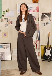 Tamnosiva oversized hoodica, bijeli uski top, široke sive hlače trenirka, smeđe tenisice, ležerni unutarnji ambijent s šarenim dekorativnim elementima.