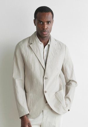 Mann trägt strukturierten hellbeigen Blazer und passende Hose, steht mit einer Hand in der Tasche vor weißem Hintergrund.