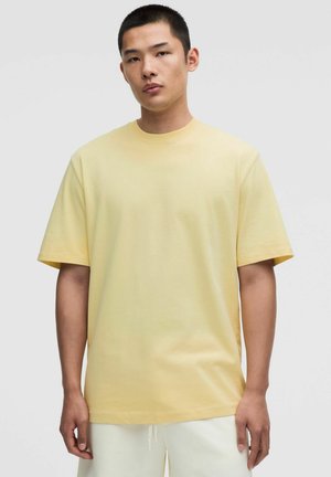 Giovane uomo in piedi che indossa una maglietta giallo chiaro a maniche corte con scollo a girocollo e pantaloni color avorio su uno sfondo grigio uniforme.