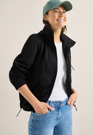 Personne souriante portant une casquette vert clair, une veste zippée noire avec des poignets élastiques, une chemise blanche et un jean bleu les mains dans les poches, faisant face à droite.