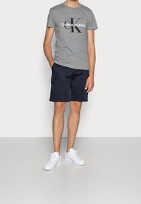 Grå bomullströja med svart "Calvin Klein Jeans"-logotyp, parat med mörkblå shorts och vita sneakers. Enfärgad bakgrund, stående pose.