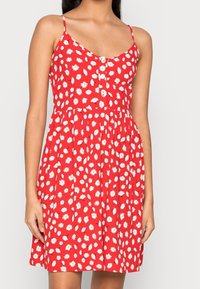 Vestido sin mangas rojas con un patrón floral de margaritas blancas, detalle de botones, silueta acampanada y tela ligera.