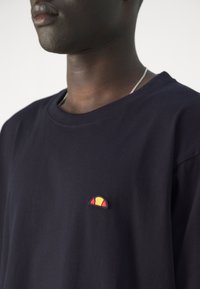 T-shirt bleu marine à manches courtes avec un col rond, comportant un petit logo brodé en rouge, jaune et noir sur la poitrine gauche.