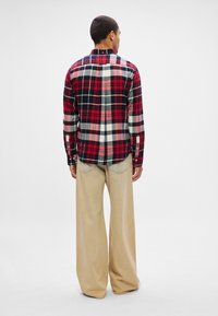 Hombre con una camisa de cuadros roja, negra y blanca de manga larga y pantalones anchos beis de pie, de espaldas, sobre un fondo blanco.
