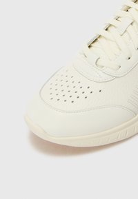 Hvit sneakers med en teksturert lær overdel, perforerte detaljer på tåen, flate snørebånd og en glatt gummisåle.