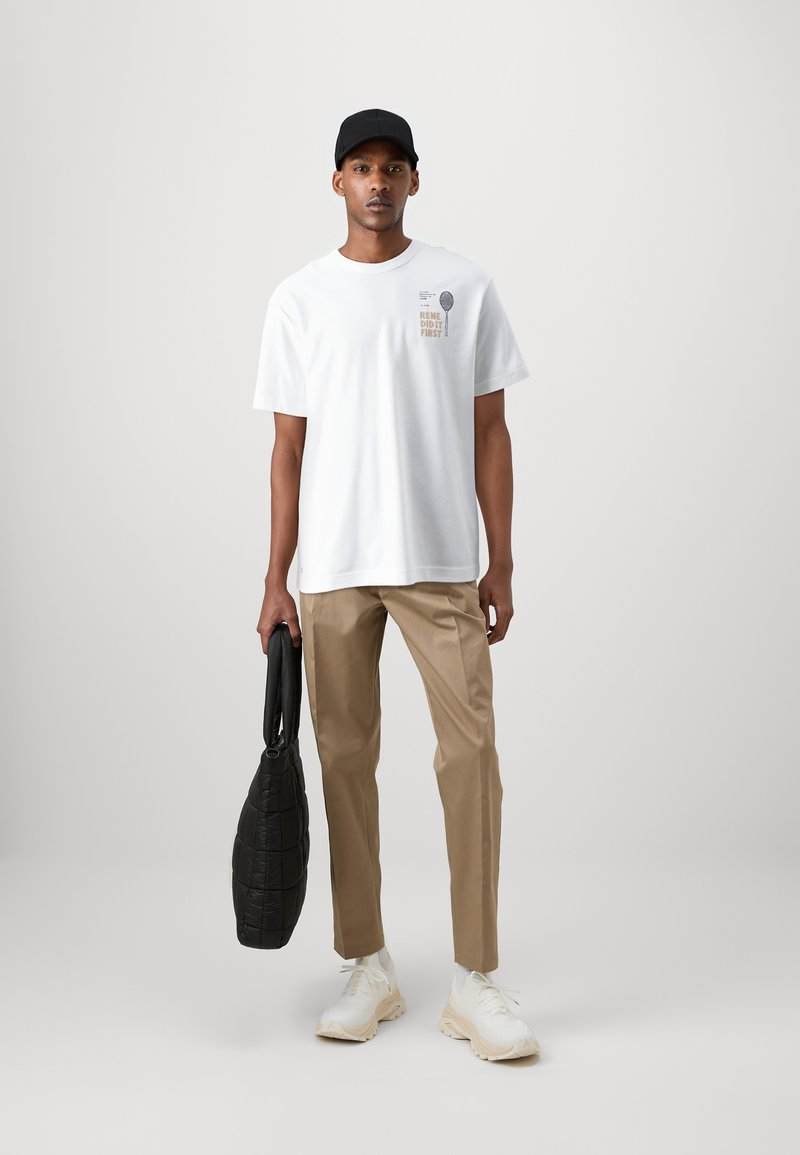 Maglietta bianca in cotone con testo stampato, pantaloni color beige, borsa nera trapuntata e sneakers color crema. Modello in piedi contro uno sfondo semplice.