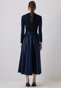 Sötétkék, hosszú ujjú ruha, testhez simuló felső résszel és bővülő szoknyával. Az anyag sima tapintású, derékpánttal kiemelve. Magas sarkú szandálok láthatók.