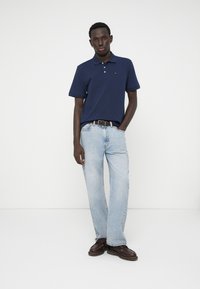 Ναυτικό μπλε πόλο μπλουζάκι με κοντά μανίκια, συνδυασμένο με ανοιχτό μπλε τζην και καφέ υποδήματα. Απλός σχεδιασμός, χαλαρή εφαρμογή και ενιαία χρώματα.