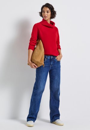 Pull à col cheminée rouge avec des détails côtelés, associé à un jean droit bleu et des baskets blanches à tige basse. Porte un sac en suede beige.