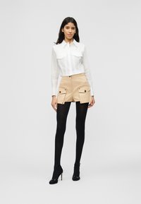 Γυναίκα με μακριά σκούρα μαλλιά που φοράει λευκό πουκάμισο με κουμπιά, μπεζ σορτς με μεγάλες τσέπες, μαύρο καλσόν και μαύρες γόβες με τακούνι.