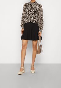 Schwarze Bluse mit Blumenmuster und langen Ärmeln, kombiniert mit einem schwarzen Rock. Cremefarbene Plateauschuhe und eine strukturiert braune Handtasche runden das Outfit ab.