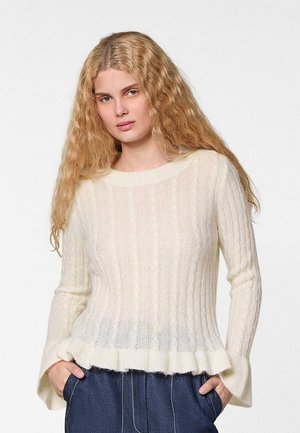 Femme aux longs cheveux blonds ondulés portant un pull blanc à torsades et un pantalon bleu, les mains dans les poches, sur fond uni.