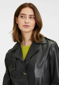 Cappotto di pelle nera con un design doppiopetto, ampie revers e due bottoni. Il modello indossa un top verde a costine sotto.