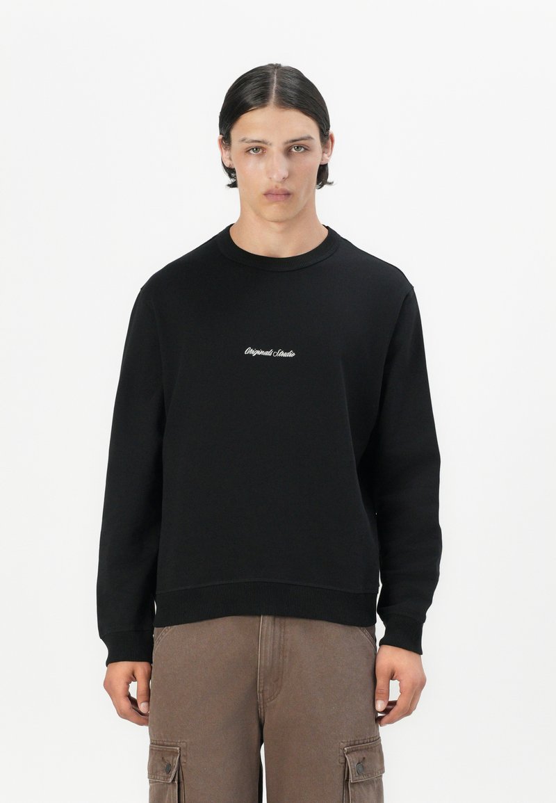Black sweatshirt with crew neckline and ribbed cuffs, featuring white embroidered text on the chest, paired with brown cargo pants.
